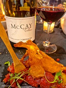 A glass of red wine, a bottle of McCay wine, and a gourmet dish with crispy fish and vegetables on a dark table.