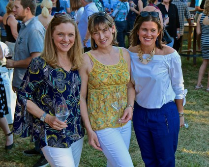 Three women smiling at an outdoor wine tasting event, holding wine glasses, with a crowd in the background.