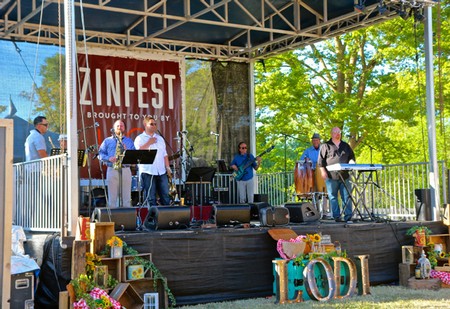 Live band performing on an outdoor stage at ZinFest festival in Lodi with colorful decorations and lush green trees.