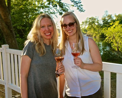 Two women enjoying wine outdoors at a Lodi Winegrape Commission event with trees and water in the background.