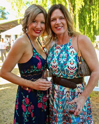 Two women smiling at an outdoor wine event, holding glasses of wine, with lush green trees in the background.