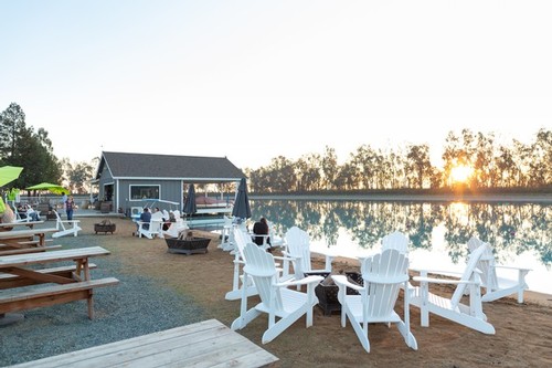 Scenic view of outdoor seating by the water with Adirondack chairs and a small building at sunset, promoting Lodi wine region.