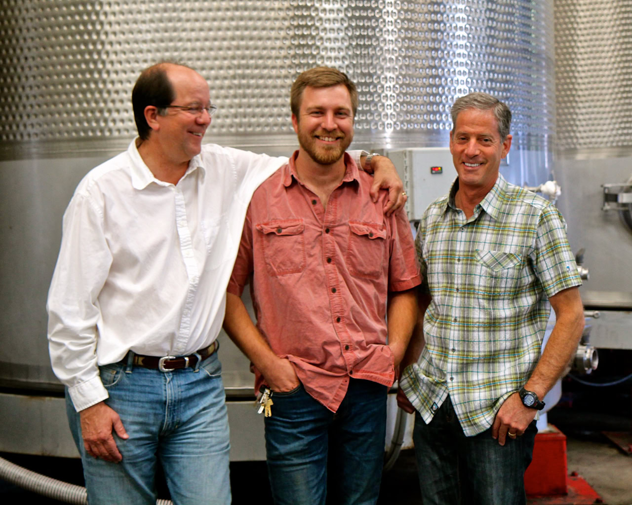 Three men smiling in a winery with stainless steel tanks in the background.