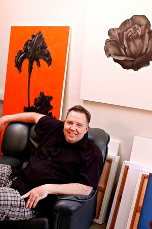 Smiling man sitting on black leather chair at Lodi Winegrape Commission art event with floral paintings in background.