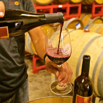 Person pouring red wine into a glass at a winery with wine barrels in the background.