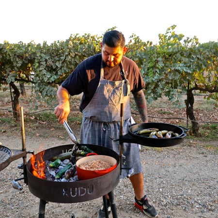 Man grilling seafood on a barbecue in a vineyard setting during sunset.