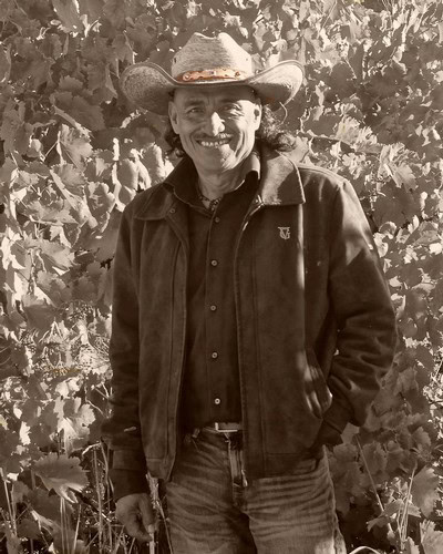 Smiling farmer in a cowboy hat standing in a vineyard with grapevines in the background.