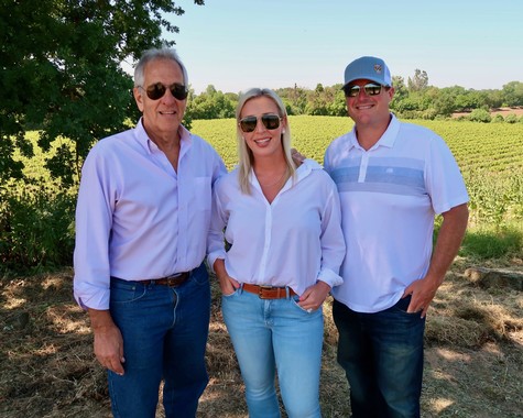 Three people standing in a vineyard, smiling, with lush grapevines and trees in the background.