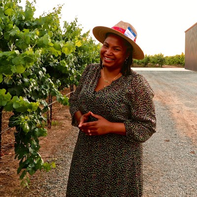 Smiling woman in a sun hat standing among grapevines at Lodi Winegrape Commission vineyard.