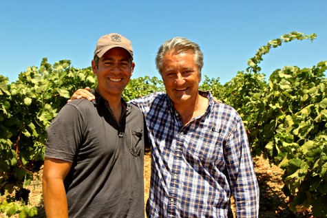 Two men smiling in a vineyard with lush grapevines under a clear blue sky.