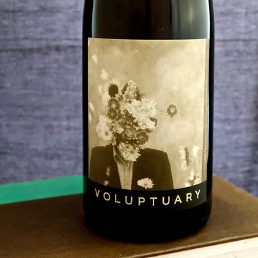 Close-up of a wine bottle featuring a surreal floral face design and the word 'Voluptuary' on the label.