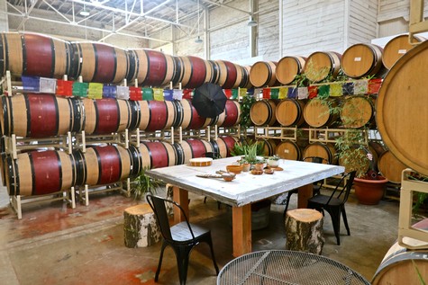Wine barrels stacked in a winery with a tasting table and chairs for visitors.