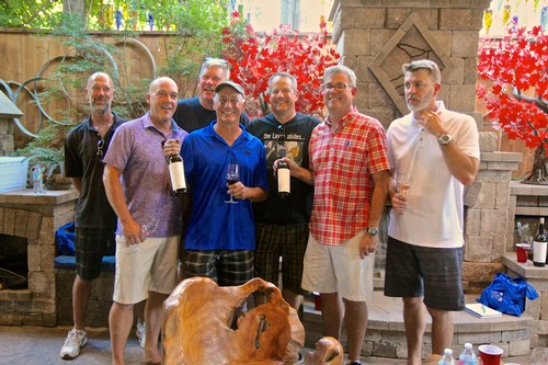 Group of men at a wine tasting event, holding wine bottles and glasses, outdoors with vibrant red foliage in the background.
