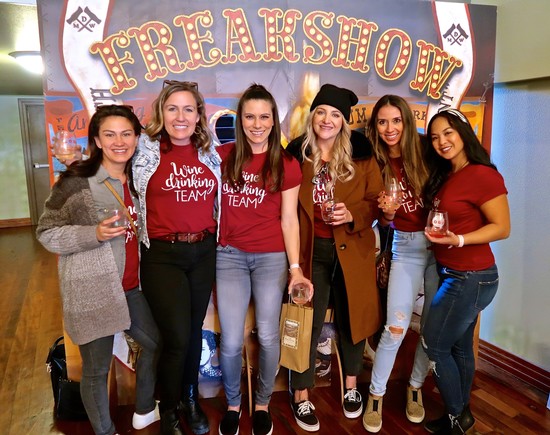 Group of women enjoying wine at the Freakshow event, smiling and holding glasses, with a colorful backdrop.