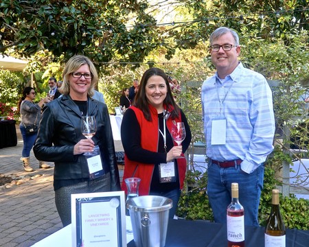 Three people at a wine tasting event holding glasses of wine outdoors with greenery in the background.