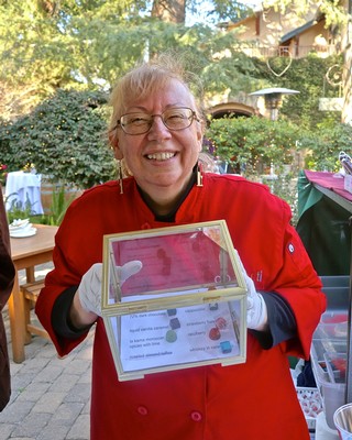 Smiling woman in red chef coat holding award at Lodi Wine event, outdoor setting with trees and tables in background.