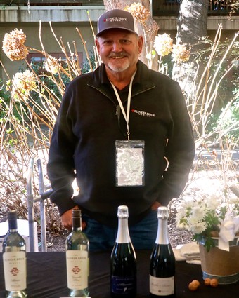 Man smiling at Lodi Winegrape Commission event with wine bottles on table.
