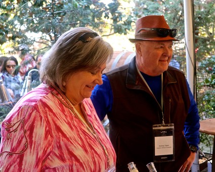 Two people at a wine event, smiling and enjoying the outdoor setting with trees in the background.