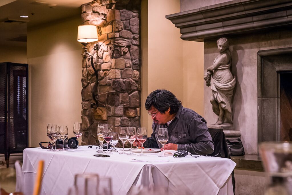 Person tasting wine at a formal event with wine glasses and a stone fireplace in the background.