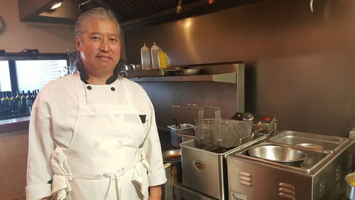 Professional chef standing in a commercial kitchen with cooking equipment and utensils.