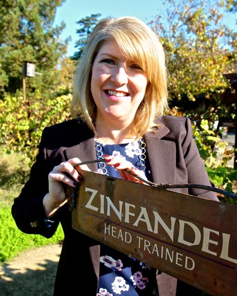 Woman smiling outdoors holding a Zinfandel head trained vineyard sign in a sunny garden setting.