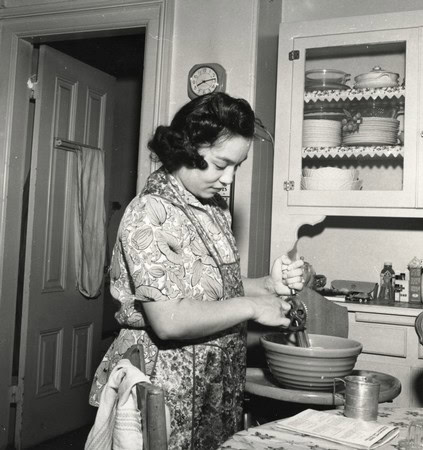 Black and white photo of a woman preparing food in a vintage kitchen, wearing a floral dress and apron, with kitchen cabinets.