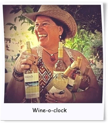 Woman holding multiple bottles of wine, smiling, wearing a straw hat, outdoors with greenery in the background.