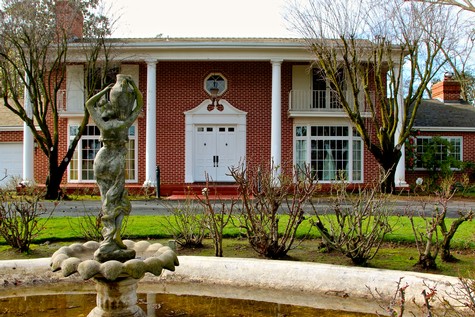 Elegant historic brick building with white columns and fountain in front, surrounded by trees and landscaped garden.