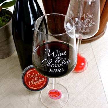 Glass of red wine with Lodi Wine & Chocolate logo, wine bottle, and chocolate on a wooden table.