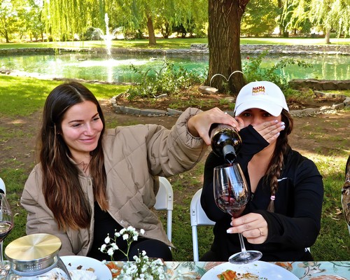 Two women enjoying wine outdoors at a Lodi Winegrape Commission event in a park with trees and a pond.