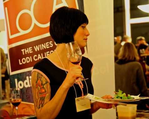 Woman tasting wine at Lodi Winegrape Commission event, holding a glass of red wine and a plate of food.