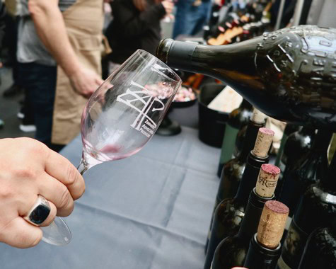Person pouring wine into a glass at a wine tasting event, with wine bottles in the background.
