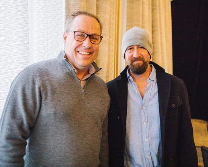 Two men smiling at a Lodi Winegrape Commission event, one wearing glasses and a gray sweater, the other in a beanie and darkj.