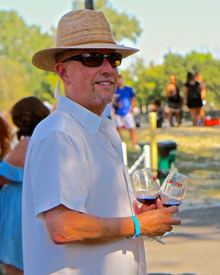 Man in straw hat and sunglasses holding wine glasses at Lodi Winegrape Commission event.