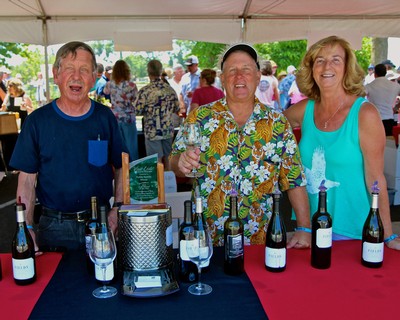 Three smiling people at a wine tasting event with bottles of wine on the table.
