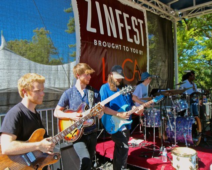 Band playing guitars and drums on stage at Lodi Wine Festival with Zinfest banner in background.