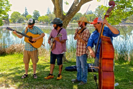 Four musicians playing instruments outdoors near a lake with trees and blue sky in the background.