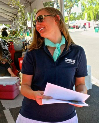 Smiling woman at Lodi Winegrape Commission event holding papers and wearing Zinfest shirt.