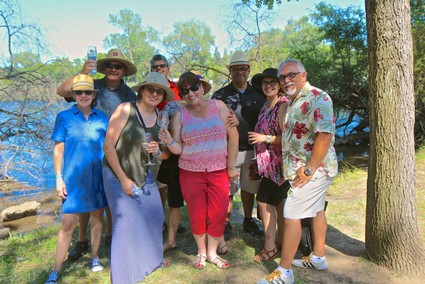 Group of people enjoying a sunny day outdoors near a river, some holding drinks, with trees and water in the background.