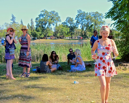 Group of people enjoying a sunny day outdoors near a pond, some sitting and some standing, with lush trees in the background.
