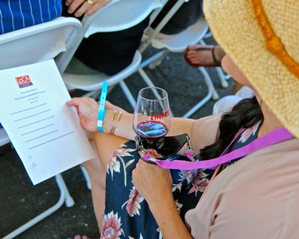 Woman with a wide-brimmed hat holding a glass of red wine and reading a program at a wine event.