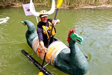 Person in a dragon boat costume paddling on water, wearing a helmet and life jacket, during a festive event.