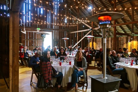 Attendees at a wine event inside a rustic barn with string lights and patio heaters.