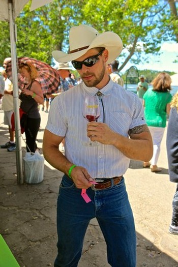 Man in cowboy hat and sunglasses enjoying wine at outdoor event with trees and crowd in background.