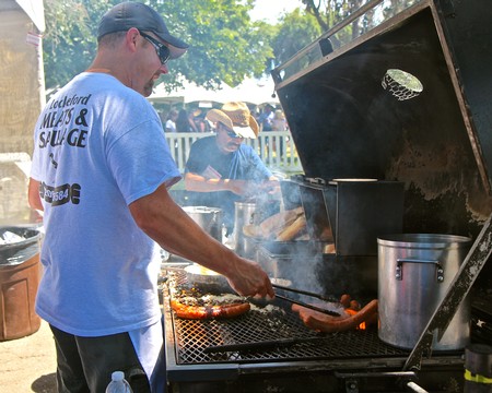 Man grilling sausages at Lodi Winegrape Commission outdoor event with people in background.