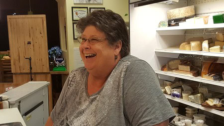 Smiling woman in a cheese shop surrounded by cheese varieties and shelves.