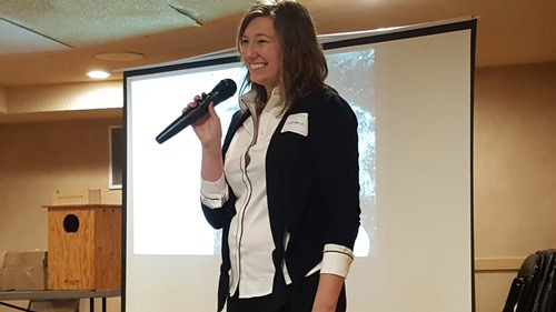 Woman speaking at Lodi Winegrape Commission event with microphone and presentation screen.